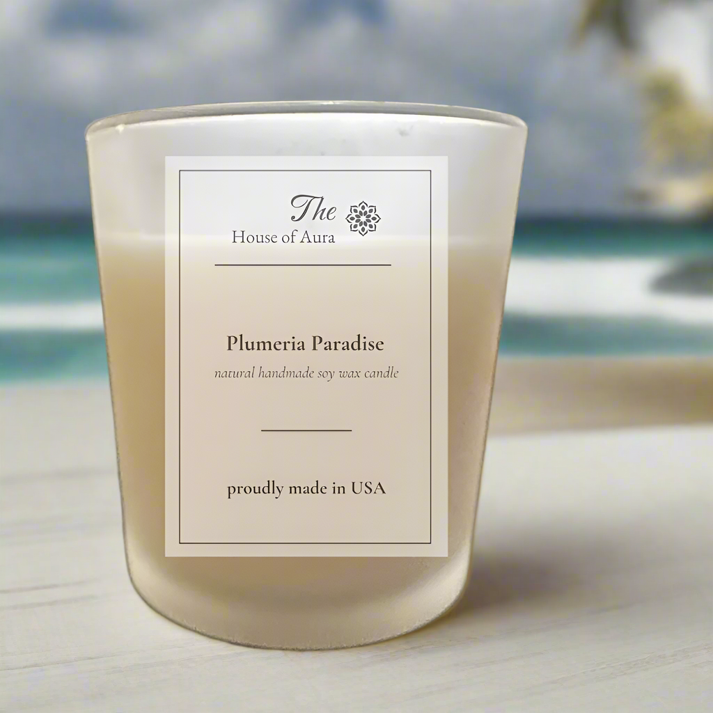 Plumeria Paradise soy wax candle in clear glass jar with tropical white and pink plumeria flowers and lush green palm leaves background, evoking a fresh island breeze scent.