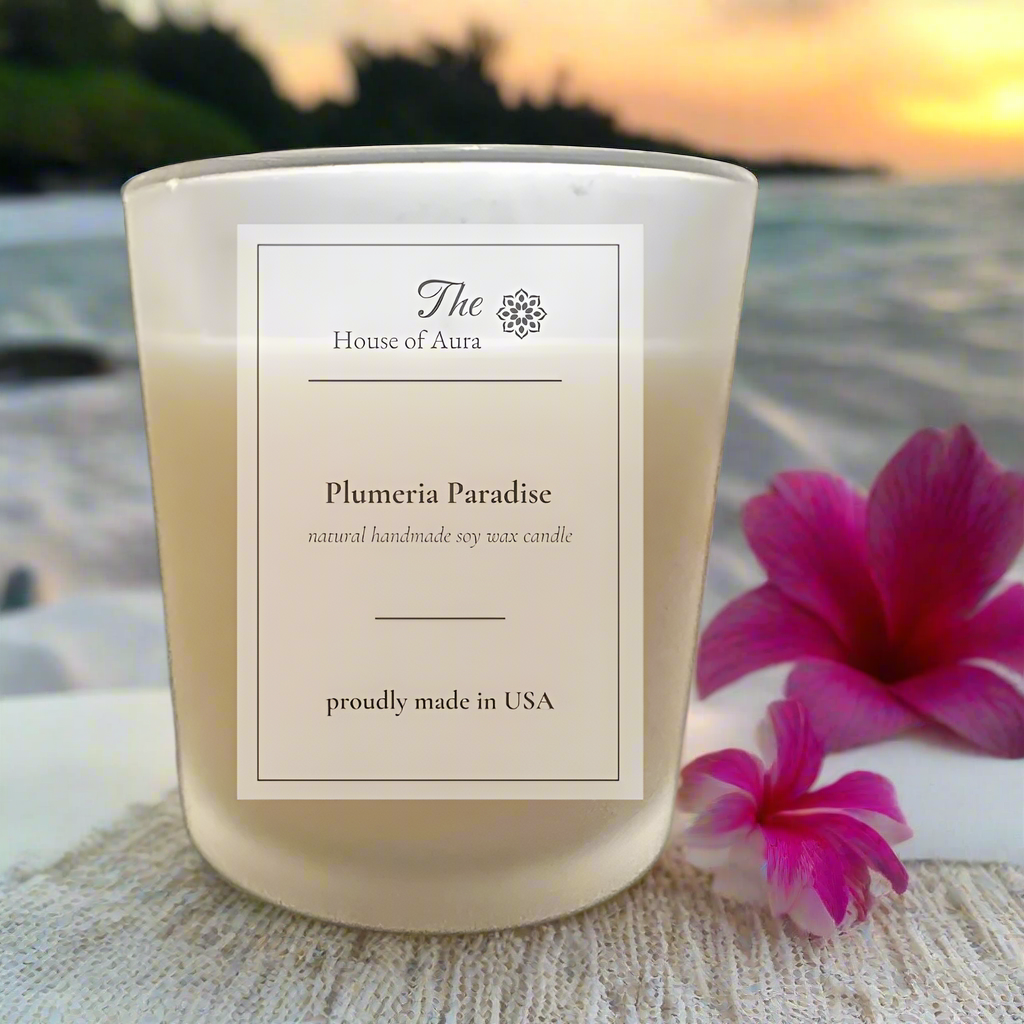 Soy wax candle labeled Plumeria Paradise set against a calm turquoise ocean sunrise with blooming white and pink plumeria flowers and soft golden light.